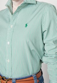 Zeleno-bíle pruhovaná košile na knoflíky s klasickým límečkem, na levé straně hrudi s malým vyšitým logem. Bavlněný materiál.