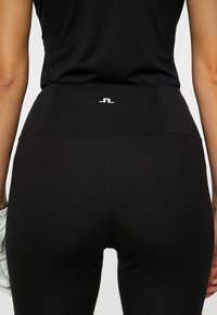 Primo piano di una persona che indossa leggings neri a vita alta e un top nero, mentre tiene un guanto bianco nella mano sinistra.