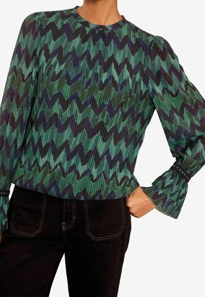 Frau trägt eine Bluse mit grünem und schwarzem Zickzackmuster, langen aufgepufften Ärmeln und schwarze Hose, Hand auf der Hüfte ruhend.