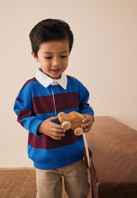 Dítě v modro-vínovém pruhovaném svetru s bílým límečkem drží dřevěné autíčko. V pozadí je dřevěný povrch.