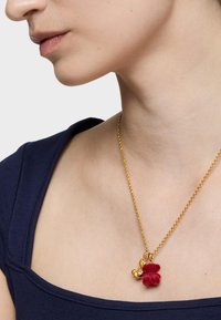 Collar de cadena de oro que presenta un pequeño charm de oso de peluche rojo con un acabado texturizado y un acento dorado. El collar se lleva sobre una blusa azul marino.