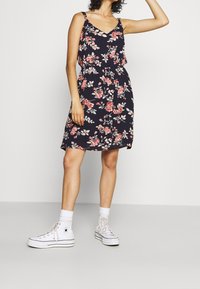 Blumenkleid für den Sommer in Marineblau mit rosa und weißen Blumen. Ärmelloses Design, tailliert und mit schwingendem Rock, kombiniert mit weißen Plateausneakern.