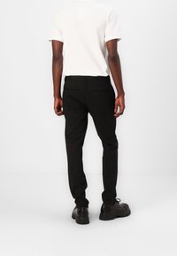 Pantalon noir à rayures avec une coupe ajustée, associé à un t-shirt blanc uni. Poches arrière visibles et chaussures noires imposantes.