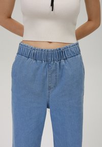 Lys blå denim bukser med elastisk liv, løs passform og subtile sømdetaljer. Brukt med en kortermet hvit ribbestrikket topp.