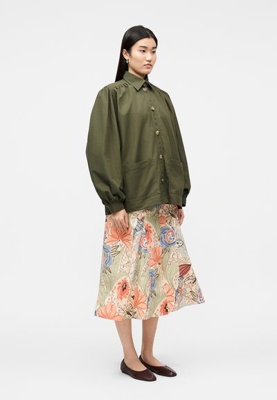 Γυναίκα στέκεται φορώντας φαρδιά πράσινη ελιά σακάκι με κουμπιά, φούστα μέσης με λουλουδάτο μοτίβο και πορτοκαλί και μπλε λεπτομέρειες, και καφέ μπαλαρίνες.
