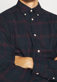 Man i mörk marinblå skjorta med knappar och tunna röda rutor, med armarna korsade över bröstet.