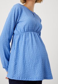 Ljust blå texturerad topp med långa ärmar, rundad halsringning och samlad midja, designad för gravidkläder.