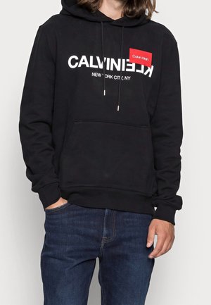 Sudadera negra con el texto blanco "CALVIN KLEIN NEW YORK CITY, NY". Presenta un bolsillo tipo canguro, cordones ajustables y puños de canalé.