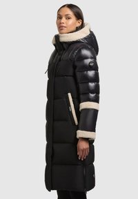 Fekete bélelt hosszú kabát magas gallérral, báránybőr díszítésekkel az ujjvégeken és a kapucnin, cipzáras zsebekkel és fényes textúrával.