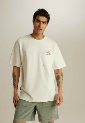 Biała bawełniana koszulka z kieszonką i pomarańczową grafiką słońca, w połączeniu z jasnorozowym szortami z kordurowej tkaniny, które mają boczne kieszenie i luźny krój.