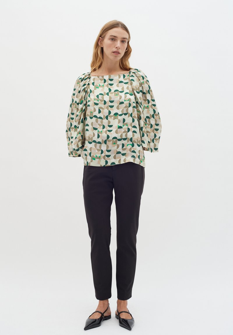 Bluză imprimată, lejeră, cu modele abstracte în verde, bej și negru; are mâneci bufante și un decolteu pătrat. Este asortată cu pantaloni strâmți negri.