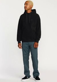 Schwarzer Kapuzenpullover mit Kordelzug, Fronttasche und Logo. Kombiniert mit taillierten, hellblauen Hosen und schwarzen Slippern.