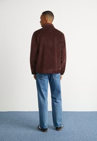 Pruunist korduroost jakk, millel on krae, nööpidega mansetid ja sirge lõikega sinised teksad, koos mustade kingadega, millel on valged triibud.