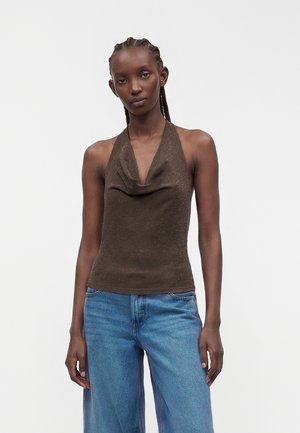Jeune femme aux cheveux foncés tressés portant un haut marron sans manches à col halter et un jean bleu, debout devant un fond clair uni.