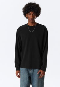 Musta, pitkähihainen t-paita, valmistettu puuvillasta, jossa pyöreä pääntie ja löysä istuvuus. Yhdistetty vaaleansinisiiin farkkuihin.