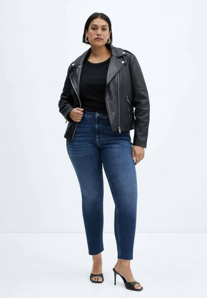 Blouson en cuir noir de motard, haut noir ajusté et jeans slim-fit bleus. La mannequin porte des talons noirs, les cheveux raides et peu d'accessoires.