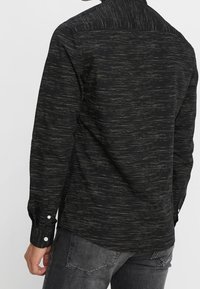 Camicia nera a maniche lunghe con tessuto testurizzato caratterizzato da sottili strisce verticali e chiusure con bottoni bianchi su polsini e colletto.