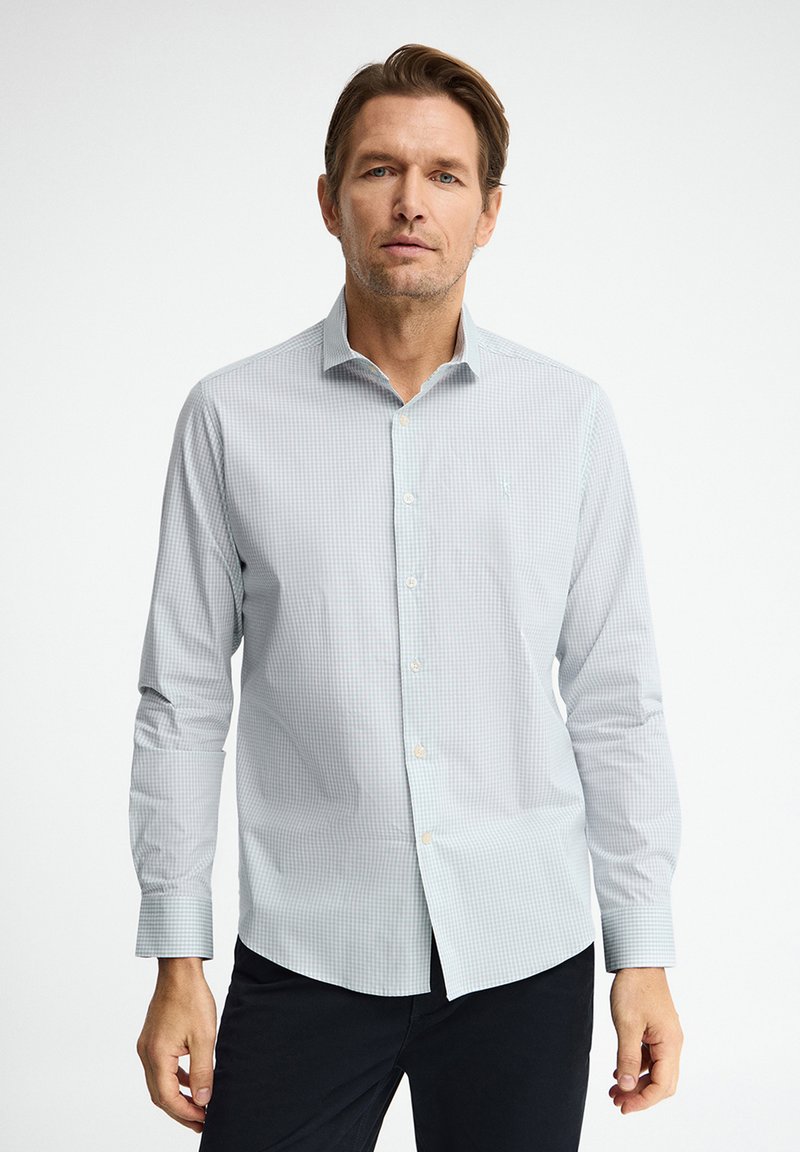 Uomo che indossa una camicia a quadretti piccoli azzurri e pantaloni scuri, in piedi contro uno sfondo bianco semplice.