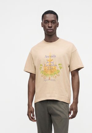 Homme portant un t-shirt beige avec un texte espagnol et un motif floral jaune, associé à un pantalon vert foncé, debout devant un fond clair uni.