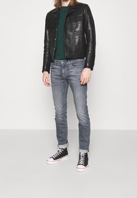 Giacca di pelle nera con due tasche sul petto, maglione verde, jeans azzurri chiari e sneakers alte nere con suole bianche e dettagli rossi.