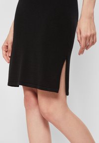 Robe en maille noire avec une fente latérale, dotée d'un ourlet droit et d'une texture subtile. Gros plan montrant une main positionnée près de l'ourlet de la robe.