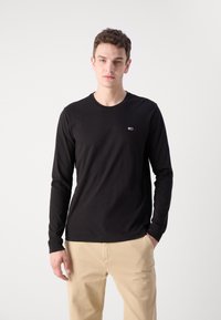 Camiseta negra de manga larga de algodón con cuello redondo, que presenta un pequeño logo bordado en el lado izquierdo del pecho. Combinada con pantalones beige.