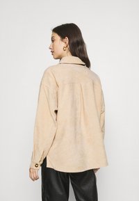 Beige korduroy košeľa s golierom, detailom na chrbte, dlhými rukávmi a gombíkovými manžetami, skombinovaná s čiernymi textúrovanými nohavicami.