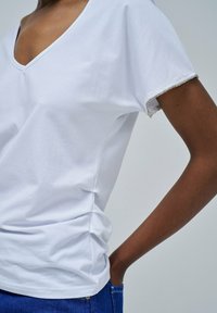 Camiseta blanca de cuello en V con mangas cortas, que presenta un borde texturizado en las mangas y un ligero fruncido a los lados. Tela de algodón suave.