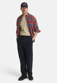Rutigt flanellskjorta i röd och blå, olivgrön t-shirt, mörka byxor och mörka skor, prydd med en marinblå keps med "GANT"-logotyp.