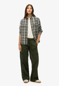Jeune femme portant une chemise à carreaux gris et vert sur un haut blanc, un pantalon cargo vert et des baskets blanches, se tenant avec une main dans la poche.
