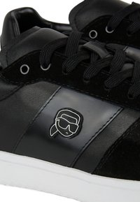 Must mustad sneaker, mis on valmistatud nahast ja sametist, silmatorkava sileda viimistluse, mustade kindade, valge kummitald ja logo embleem külgedel.