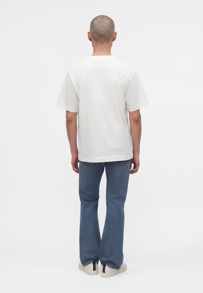 Homme à la tête rasée, debout de dos, portant un t-shirt blanc uni, un jean bleu et des baskets blanches avec des bandes noires et vertes.