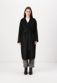 Abrigo de lana negro con cintura ceñida, mangas largas y dos bolsillos frontales. Llevado sobre pantalones de estampado y botas de tobillo.
