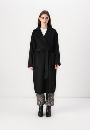 Manteau en laine noir avec une taille ceinturée, manches longues et deux poches avant. Porté sur un pantalon à motifs et des bottines.