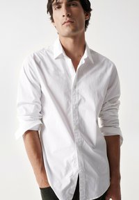Camisa blanca de botones hecha de algodón. Presenta mangas largas arremangadas hasta los codos, un cuello clásico y un dobladillo recto. Diseño minimalista.