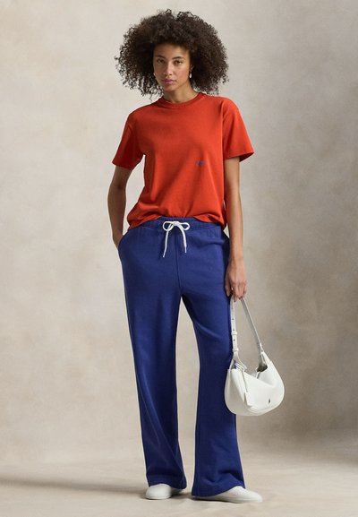 Κόκκινο βαμβακερό T-shirt με κοντά μανίκια και μικρό μπλε λογότυπο, σε συνδυασμό με μπλε φόρμα με κορδόνι και λευκή τσάντα χειρός.