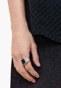 Sølvring med rektangulært design og en sort mat firkantet overflade, båret på en hånd mod en mørk stofbaggrund.