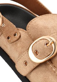 Sandalia de ante beige con punta redondeada, correas ajustables dobles con herrajes en tono dorado y suela de goma negra. Textura suave y diseño casual.