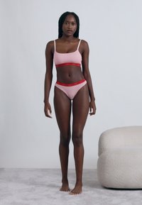 Ružová vzorovaná braletka a zodpovedajúce bikiny s červenou elastickou páskou. Hladká textúra a priliehavý strih. Jednoduché pozadie a mäkké biele kreslo.