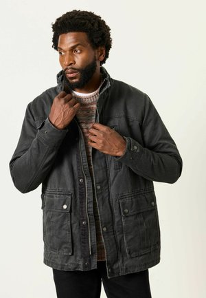 Mann, der den Kragen einer dunkelgrauen Jacke über einem mehrfarbigen Strickpullover richtet, stehend vor einem einfarbigen hellen Hintergrund.