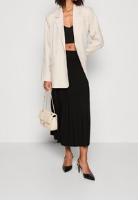 Blazer di lino beige, top corto nero, gonna plissettata nera, scarpe con tacco nero, e una borsa crema texturizzata con tracolla a catena.