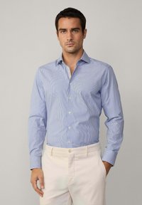 Camicia a righe blu chiaro con maniche lunghe, design con bottoni e bottoni bianchi, abbinata a pantaloni chiari.