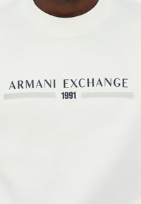 Fehér pulóver, puha anyagból készült, kék betűkkel "ARMANI EXCHANGE" felirattal és alatta "1991" számjeggyel, szürke díszítő vonalakkal.