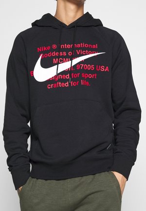 Osoba ubrana w czarną bluzę Nike z dużym białym logo i czerwonym napisem, ręce w kieszeniach, w połączeniu z oliwkowozielonymi spodniami.