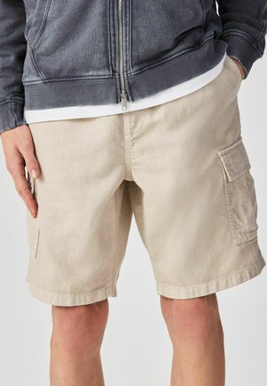 Mann trägt beige Cargo-Shorts und eine dunkelgraue Jacke mit Reißverschluss, die Hände in den Taschen, gezeigt von der Mitte des Oberschenkels bis zur Taille.