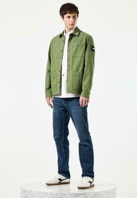 Lysgrøn casual jakke med knaplukning, to brystlommer, båret over en hvid skjorte, parret med blå jeans og hvide sneakers.