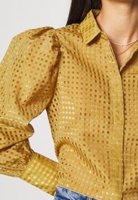 Blouse jaune à carreaux en tissu transparent, manches longues bouffantes, col structuré et accents métalliques subtils dans le motif.