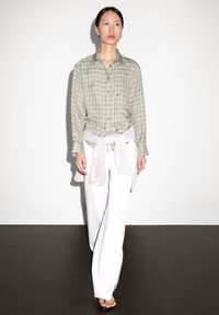 Groene geruite blouse met lange mouwen, gedragen over een lichtroze trui die om de taille is geknoopt, in combinatie met witte wijde broek.