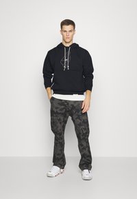 Felpa nera con grafica bianca, pantaloni cargo grigi a camouflage e scarpe da ginnastica bianche. La felpa presenta dei lacci; i pantaloni cargo hanno molteplici tasche.