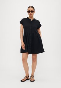Rochie neagră cu nasturi, cu mâneci scurte și fustă în straturi. Fabricată dintr-un material ușor, cu un croi lejer. Asortată cu sandale negre cu curele.